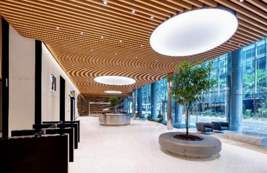 Spacious modern lobby with wooden slat ceiling, large circular lights, indoor trees, floor-to-ceiling windows, and seating areas.
