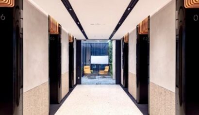 A modern lift lobby with six black lift doors, white walls, and a view of two yellow chairs and a glass wall at the far end.
