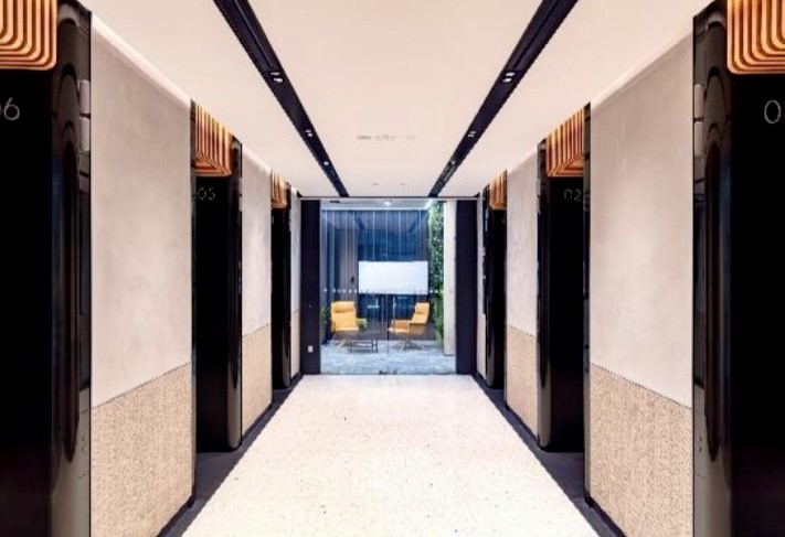 A modern lift lobby with six black lift doors, white walls, and a view of two yellow chairs and a glass wall at the far end.