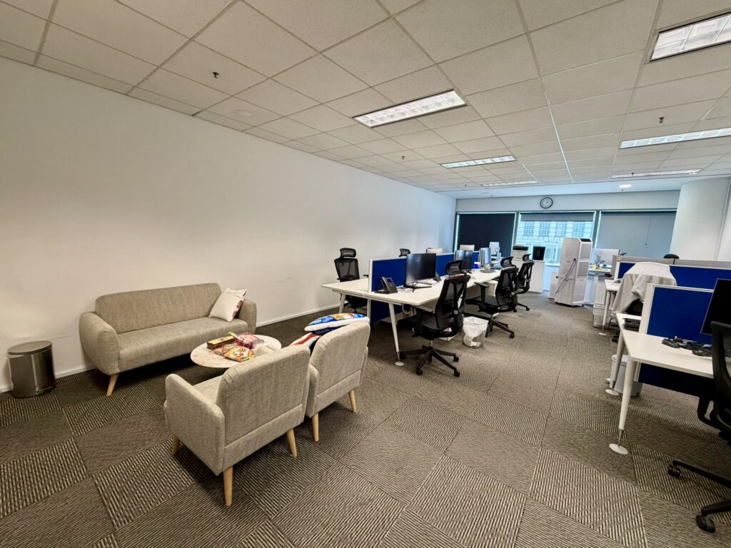 Modern office space with desks, computers, and chairs. A lounge area with a sofa and armchairs is in the foreground. The office appears clean and organised.
