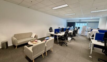 Modern office space with desks, computers, and chairs. A lounge area with a sofa and armchairs is in the foreground. The office appears clean and organised.