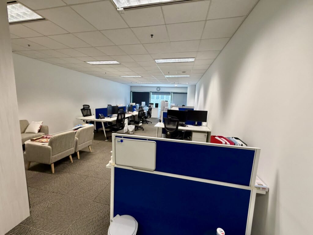 Modern office space with several desks, computers, office chairs, and sofas; overhead lighting and a window at the far end of the room.