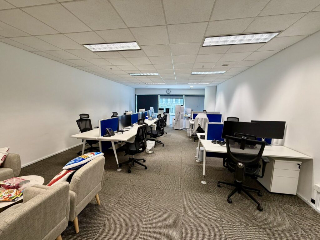 A modern office space with several empty desks and office chairs, computers on desks, and a sofa with cushions along the left wall. Fluorescent ceiling lights are on.