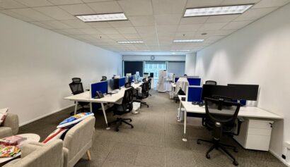 A modern office space with several empty desks and office chairs, computers on desks, and a sofa with cushions along the left wall. Fluorescent ceiling lights are on.