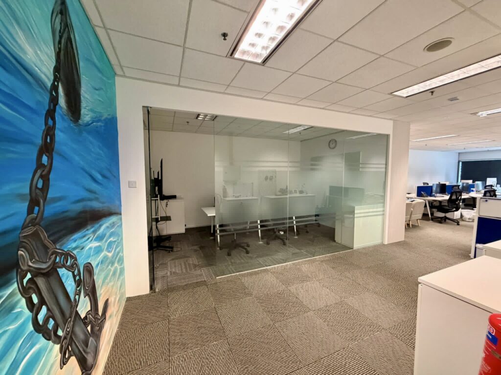 Modern office space with glass-walled meeting room, desk workstations, and a large wall mural of a chain and anchor in water on the left. The area is well-lit with overhead lights.