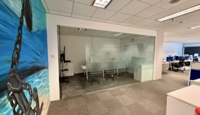 Modern office space with glass-walled meeting room, desk workstations, and a large wall mural of a chain and anchor in water on the left. The area is well-lit with overhead lights.