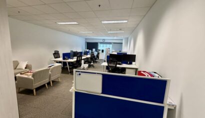 Modern office space with several desks, computers, office chairs, and sofas; overhead lighting and a window at the far end of the room.