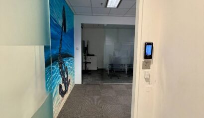 Office doorway with a glass door, a wall mural of an anchor by the sea on the left, and a view into a room with office furniture and a whiteboard in the background.