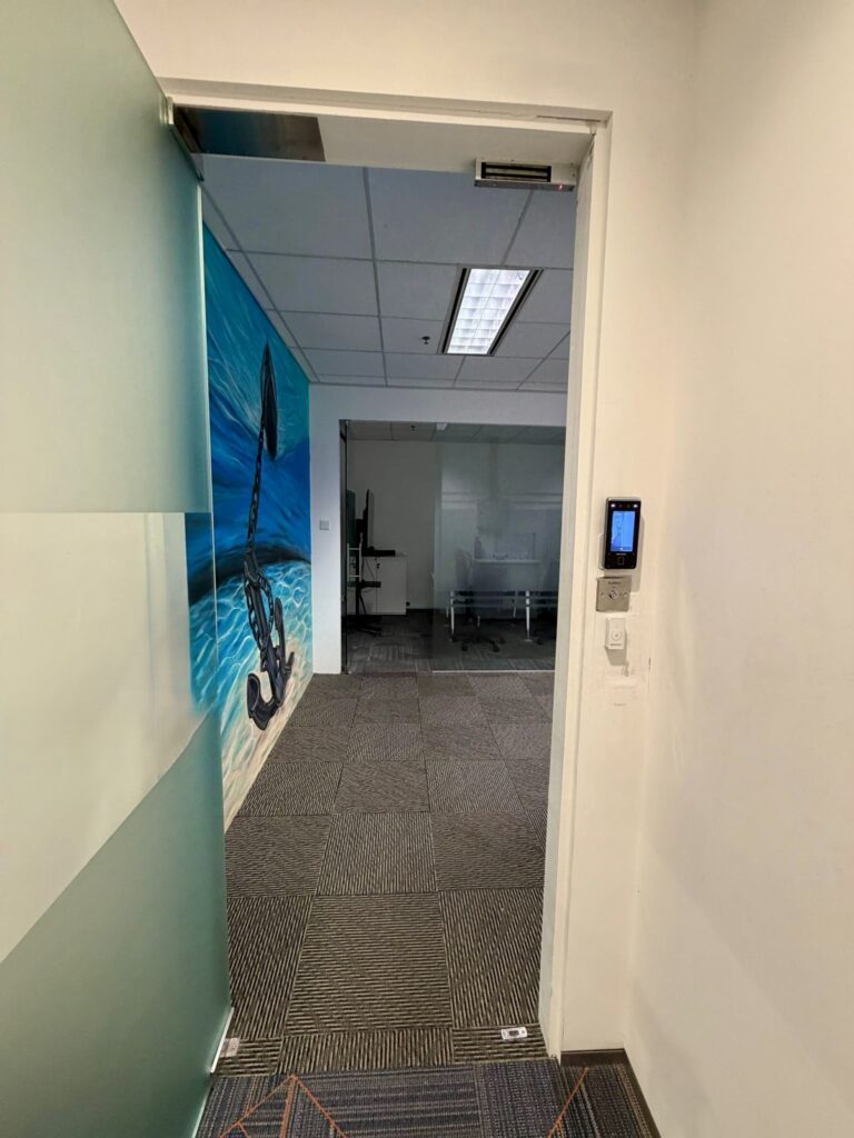 Office doorway with a glass door, a wall mural of an anchor by the sea on the left, and a view into a room with office furniture and a whiteboard in the background.