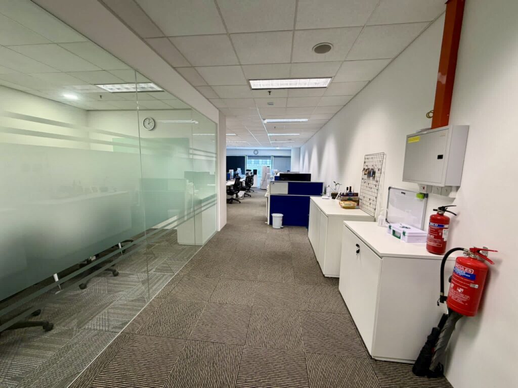 A modern office corridor with glass-walled meeting rooms on the left and white cupboards, a fire extinguisher, and a first aid kit on the right; workstations are visible ahead.
