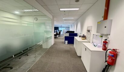 A modern office corridor with glass-walled meeting rooms on the left and white cupboards, a fire extinguisher, and a first aid kit on the right; workstations are visible ahead.