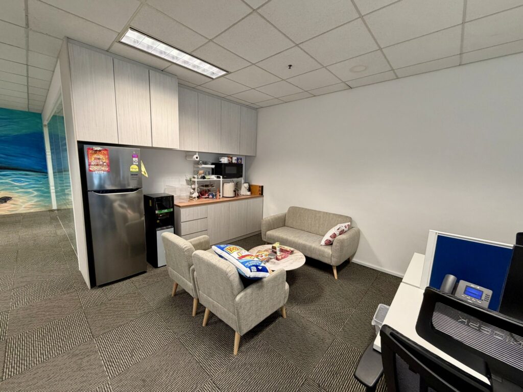 Office break area with a kitchenette, small fridge, coffee machine, two sofas, armchairs, a round table, and a worktop in the foreground.