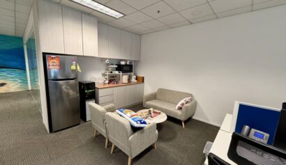 Office break area with a kitchenette, small fridge, coffee machine, two sofas, armchairs, a round table, and a worktop in the foreground.