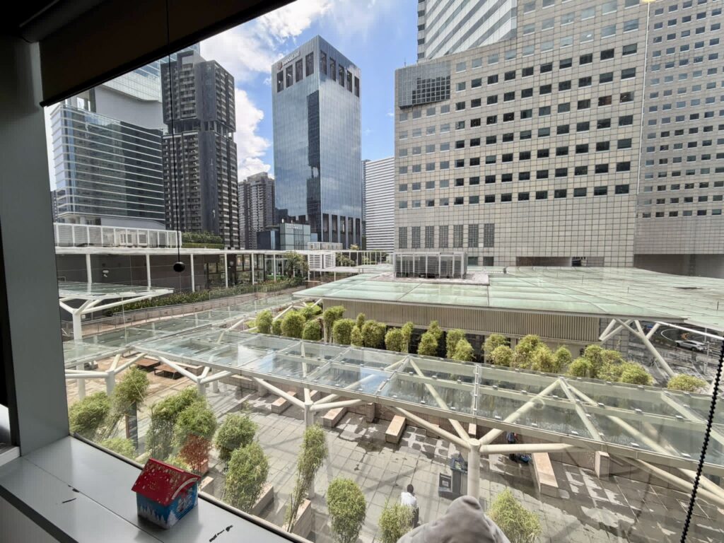 View from a window overlooking a modern cityscape with tall glass buildings, green trees, and a covered walkway in an urban plaza. A red toy house sits on the window sill.