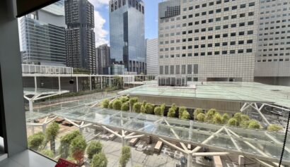 View from a window overlooking a modern cityscape with tall glass buildings, green trees, and a covered walkway in an urban plaza. A red toy house sits on the window sill.