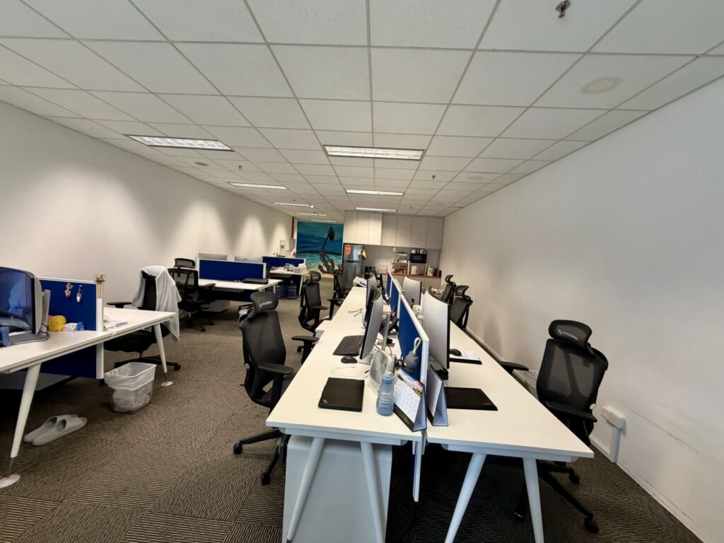 Open-plan office with several empty desks, computer monitors, office chairs, and a kitchenette in the background. The space is brightly lit with white walls and ceiling tiles.