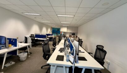 Open-plan office with several empty desks, computer monitors, office chairs, and a kitchenette in the background. The space is brightly lit with white walls and ceiling tiles.
