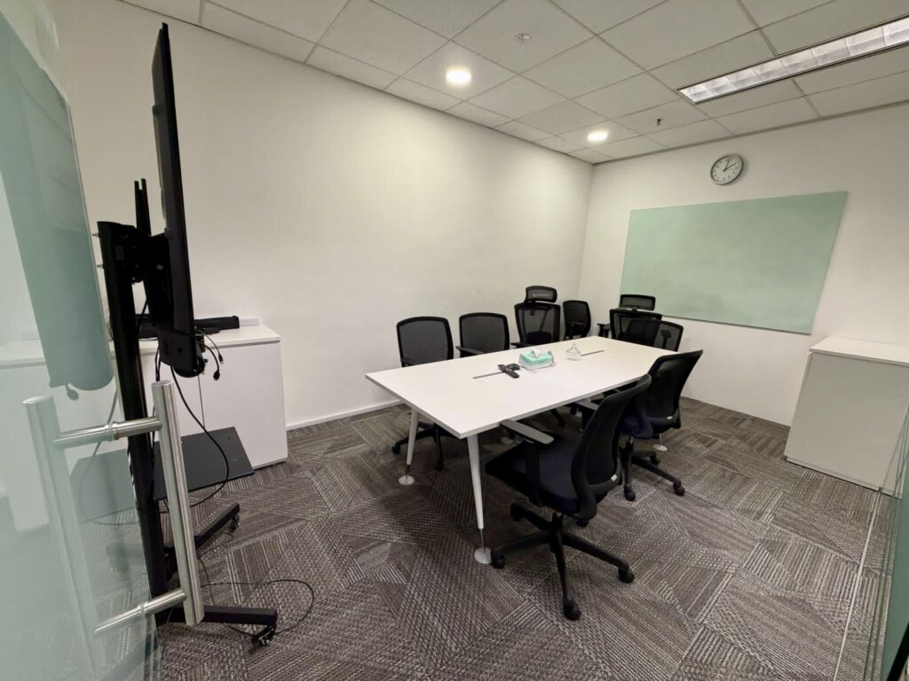 A conference room with six chairs around a white table, a wall clock, a large screen, a whiteboard, and office supplies on the table.