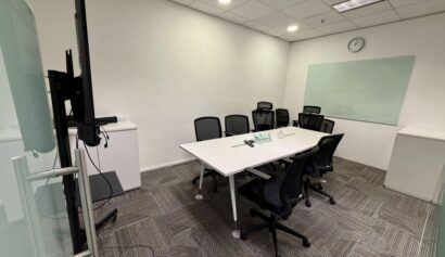 A conference room with six chairs around a white table, a wall clock, a large screen, a whiteboard, and office supplies on the table.
