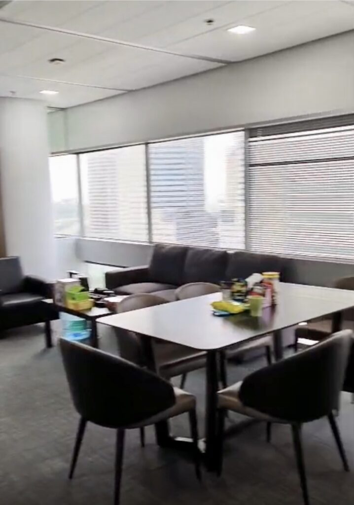 Office break room with a table, four chairs, a sofa, armchair, and snacks on the table. Large windows with blinds and city buildings visible outside.