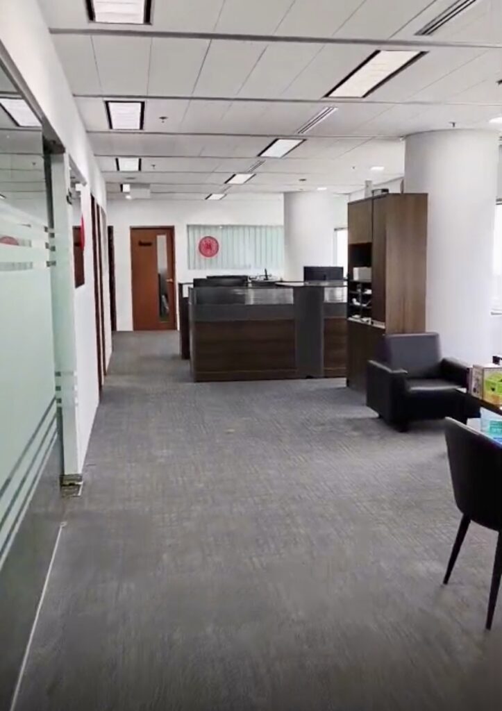 Modern office corridor with grey carpet, glass walls on the left, dark wood reception desk at the end, black armchairs, and shelves on the right. Bright overhead lights illuminate the space.