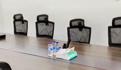 A modern conference room with a long table, six office chairs, water bottles, tissues, and office supplies arranged at the centre.