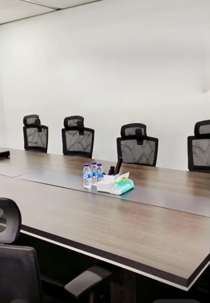 A modern conference room with a long table, six office chairs, water bottles, tissues, and office supplies arranged at the centre.