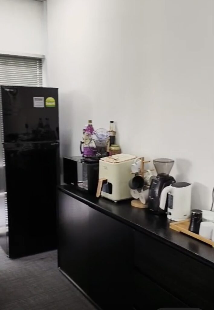 Office kitchenette with a black fridge, countertop appliances including a coffee machine, toaster, kettle, mugs, and bottles against a white wall.