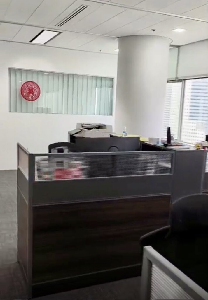 Modern office reception area with a desk, computer, office supplies, and a window. A red decorative plate hangs on a white wall behind glass panels.