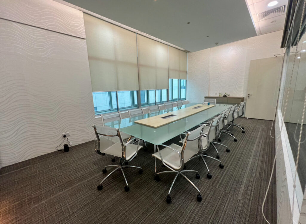 Modern conference room with a long glass table, ten white chairs, window blinds, wall panels, and a worktop with a sink in the corner.