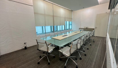 Modern conference room with a long glass table, ten white chairs, window blinds, wall panels, and a worktop with a sink in the corner.