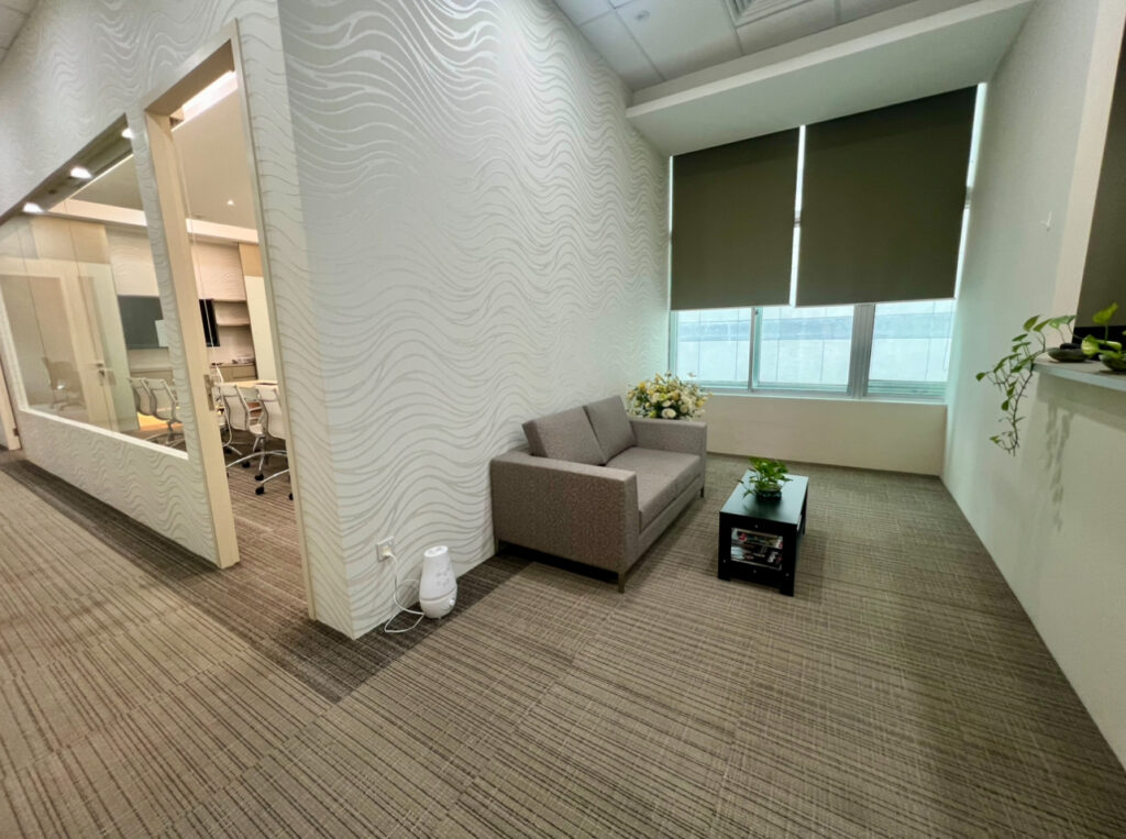 Small office waiting area with a grey sofa, coffee table, potted plants, closed blinds, and a visible meeting room through glass walls on the left.