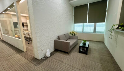 Small office waiting area with a grey sofa, coffee table, potted plants, closed blinds, and a visible meeting room through glass walls on the left.