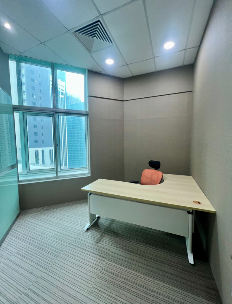 Small, modern office with a single desk, an orange office chair, large window, beige walls, and striped carpet. Tall buildings are visible outside the window.