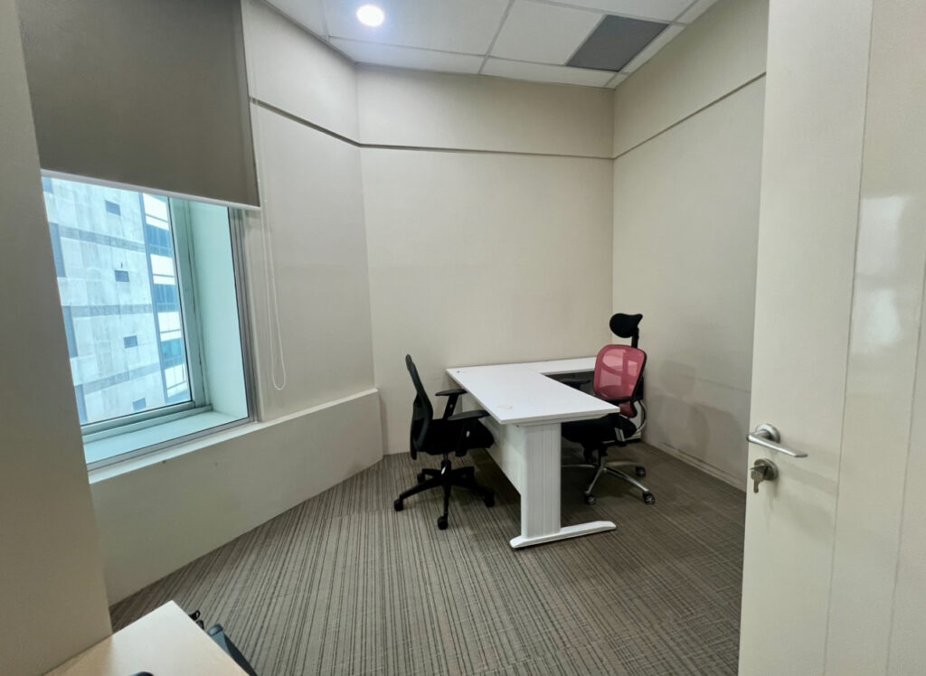 Small office room with two chairs and a white desk near a window, beige walls, carpeted floor, and overhead lighting.