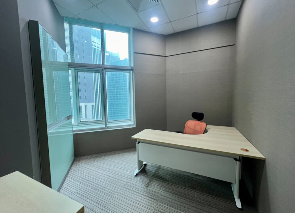 A small office with a single desk and chair near a large window overlooking city buildings. The space has neutral walls and carpeted flooring.