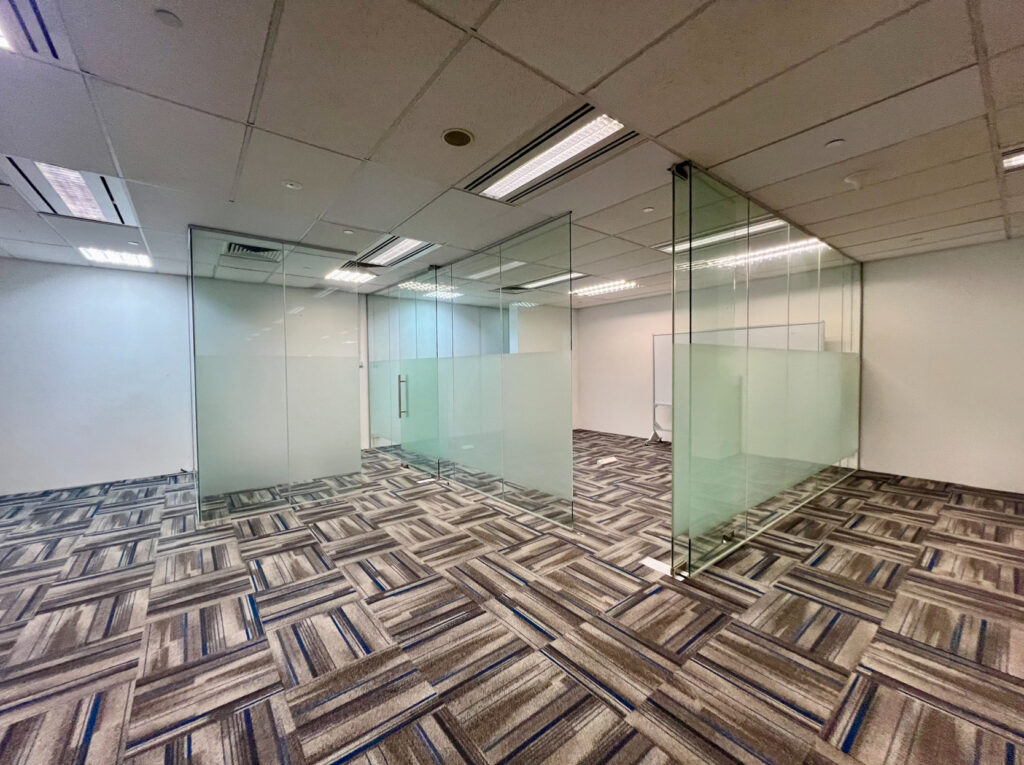 Empty office space with glass-walled meeting rooms, patterned carpet, and white ceiling tiles with recessed lighting.