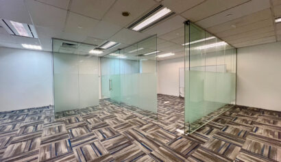 Empty office space with glass-walled meeting rooms, patterned carpet, and white ceiling tiles with recessed lighting.