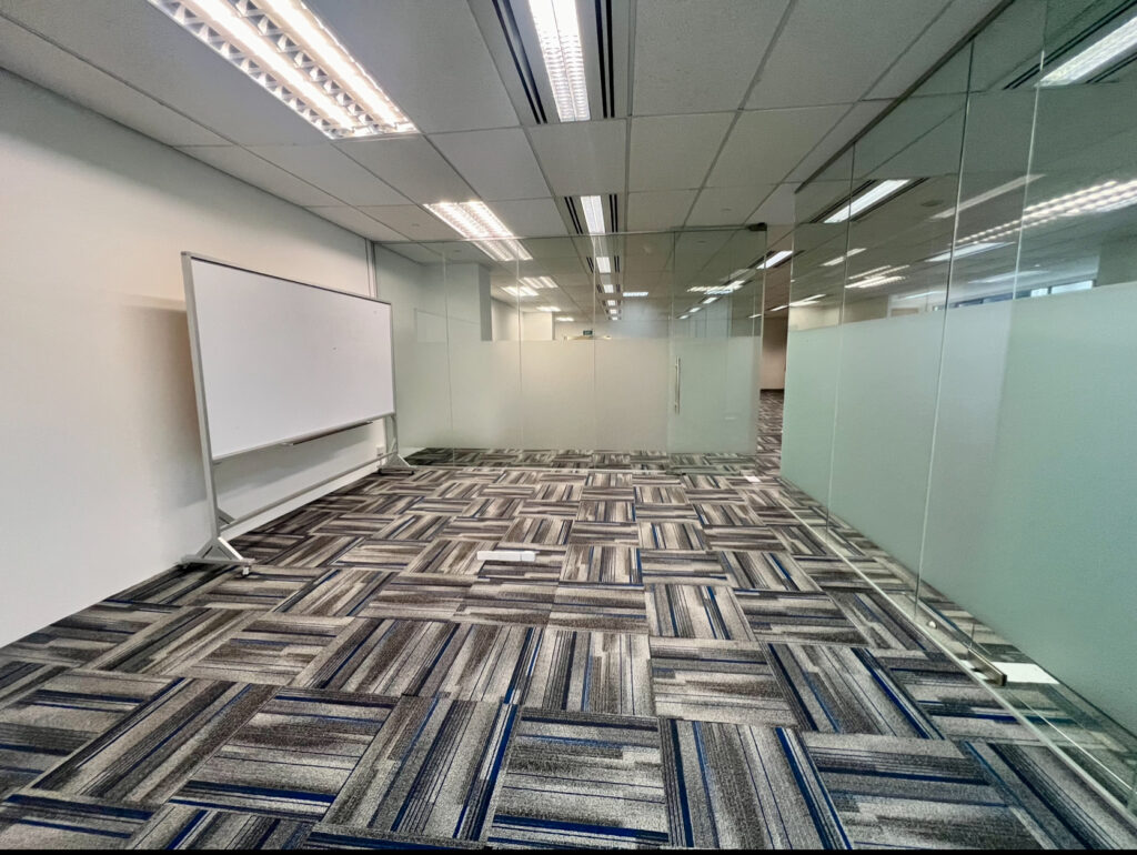 Empty office space with glass walls, patterned carpet, overhead lights, and a blank whiteboard on the left side of the room.
