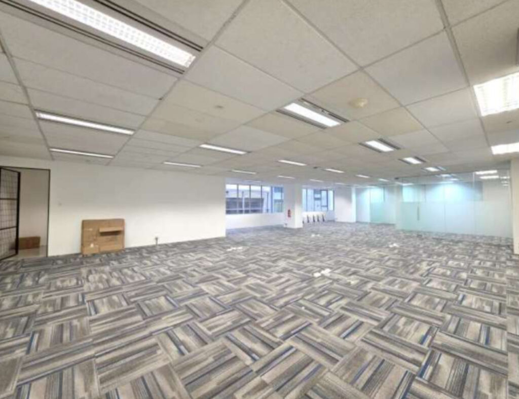 Empty office space with patterned carpet, white walls, fluorescent ceiling lights, and large windows along the far wall. A small cardboard box is on the left side of the room.