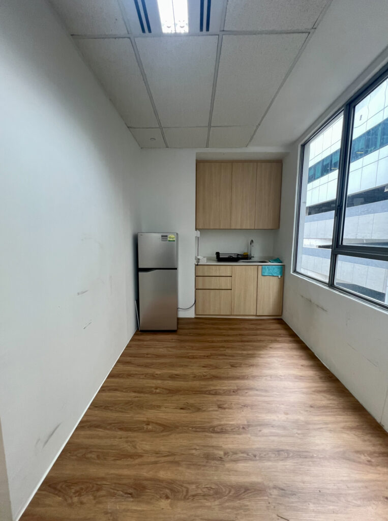 Small kitchen with wooden flooring, a stainless steel fridge, upper and lower cupboards, a sink, and a window letting in natural light.