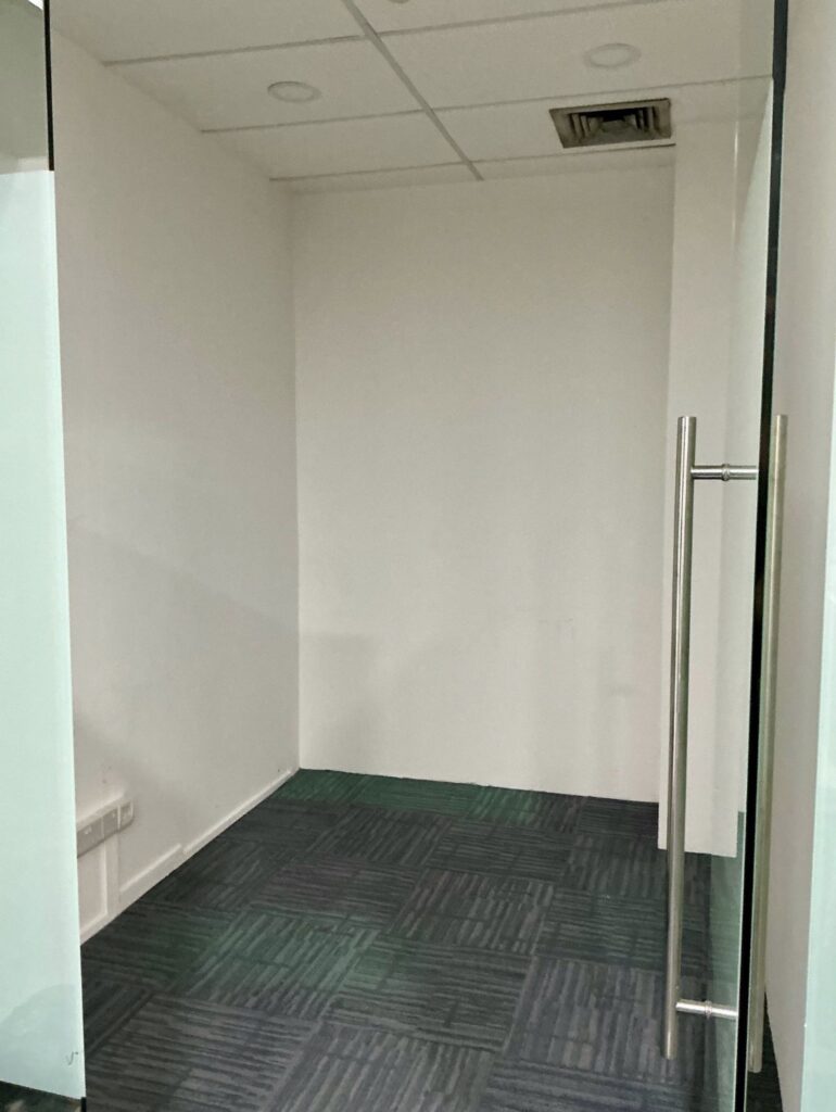 Small empty office space with white walls, green patterned carpet tiles, a glass door with a metal handle, and a ceiling vent visible.