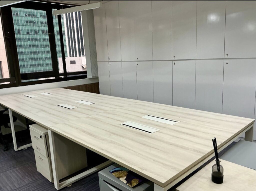 Empty modern office with a large light wood conference table, white cupboards in the background, and a window overlooking city buildings. A diffuser is on the corner of the table.
