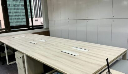Empty modern office with a large light wood conference table, white cupboards in the background, and a window overlooking city buildings. A diffuser is on the corner of the table.