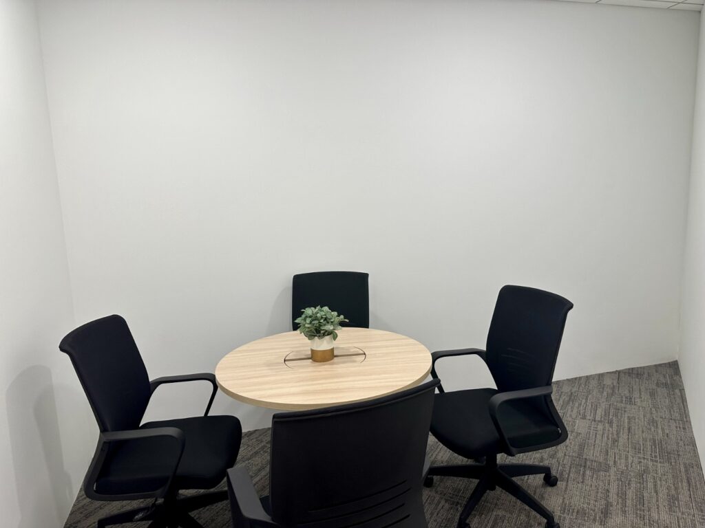 Small meeting room with a round wooden table, four black chairs, a potted plant in the centre, and plain white walls.