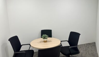Small meeting room with a round wooden table, four black chairs, a potted plant in the centre, and plain white walls.