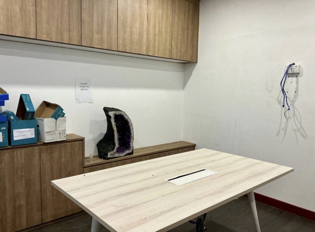 A small office room with a light wood table, cupboards, boxes, a large amethyst geode, a wall socket with cables, and a paper sign taped to the wall.