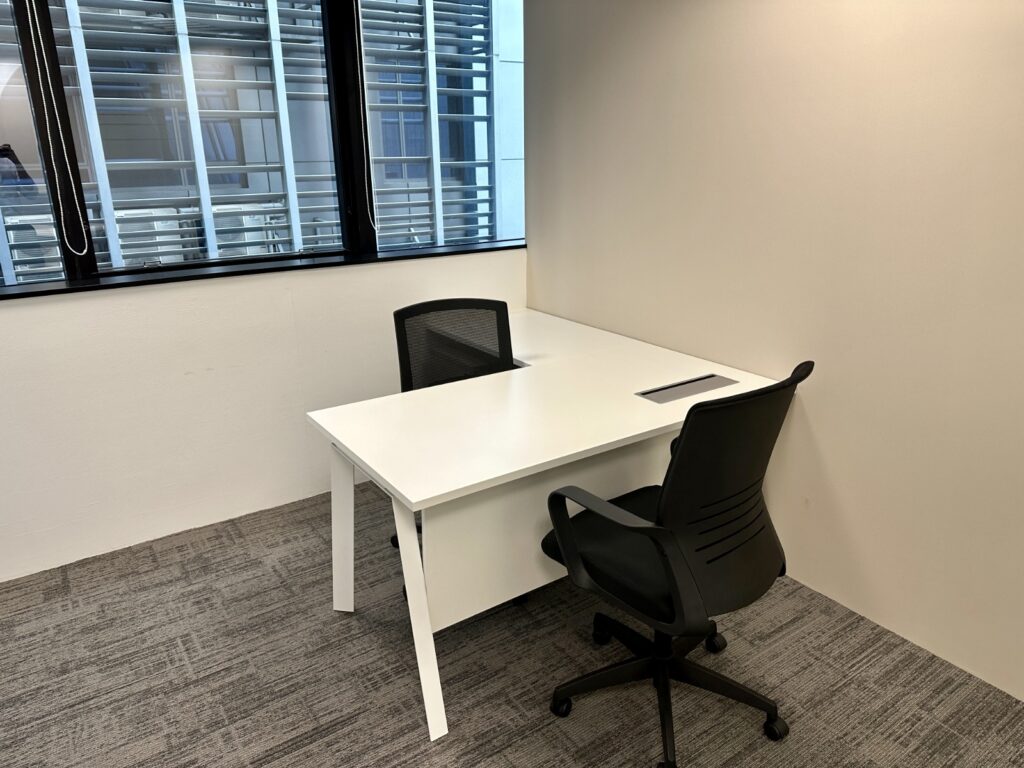 A small office with two black chairs and a white desk near a window with blinds, set against beige walls and a grey carpeted floor.