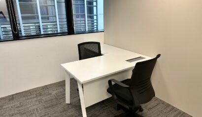A small office with two black chairs and a white desk near a window with blinds, set against beige walls and a grey carpeted floor.