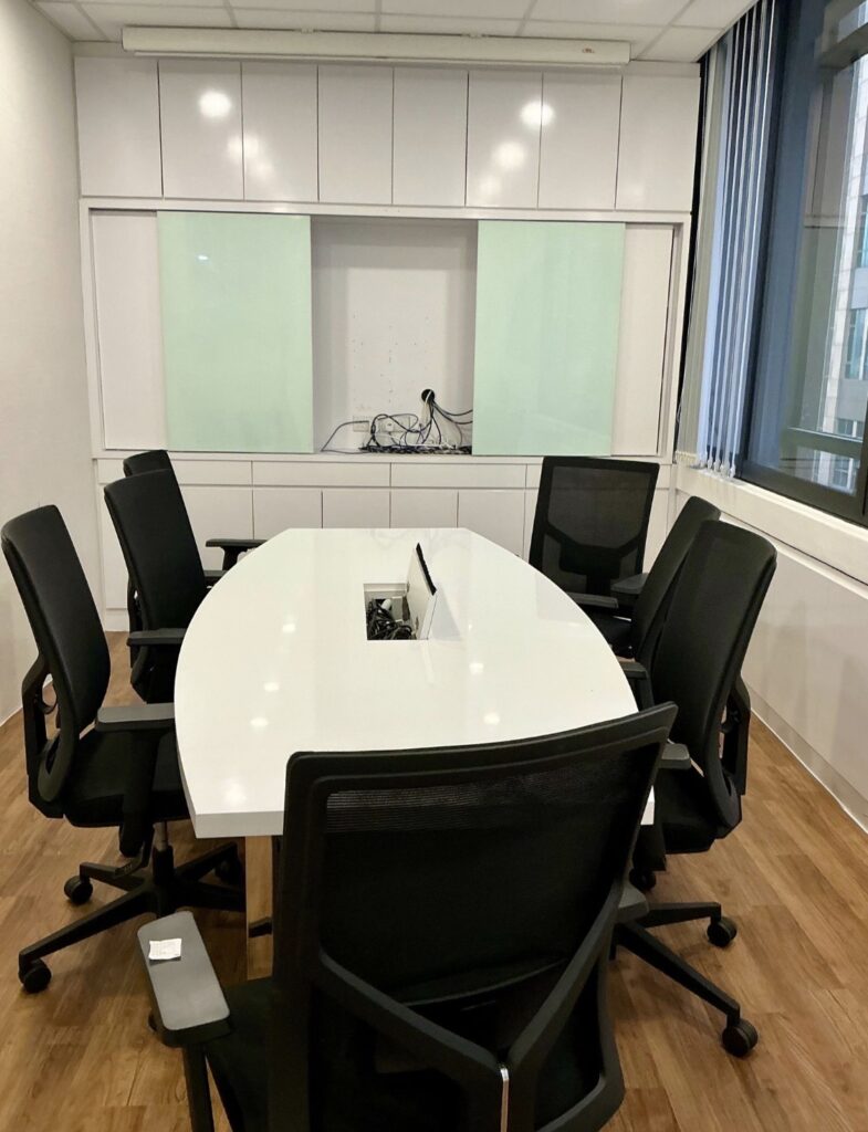 A modern conference room with a white oval table, six black office chairs, wooden flooring, and a wall cupboard with exposed wires.